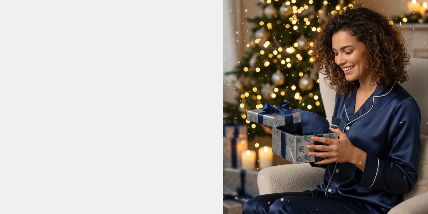 Vrouw in satijnen pyjama opent luxe PurelyGoods cadeau met blauw slaapmasker voor een kerstboom, sfeervolle kerstverlichting op de achtergrond.