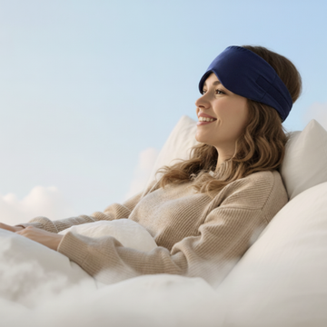 Vrouw ligt ontspannen in bed met donkerblauw PurelyGoods slaapmasker, glimlachend in ochtendlicht, ideaal voor rust en verduistering.