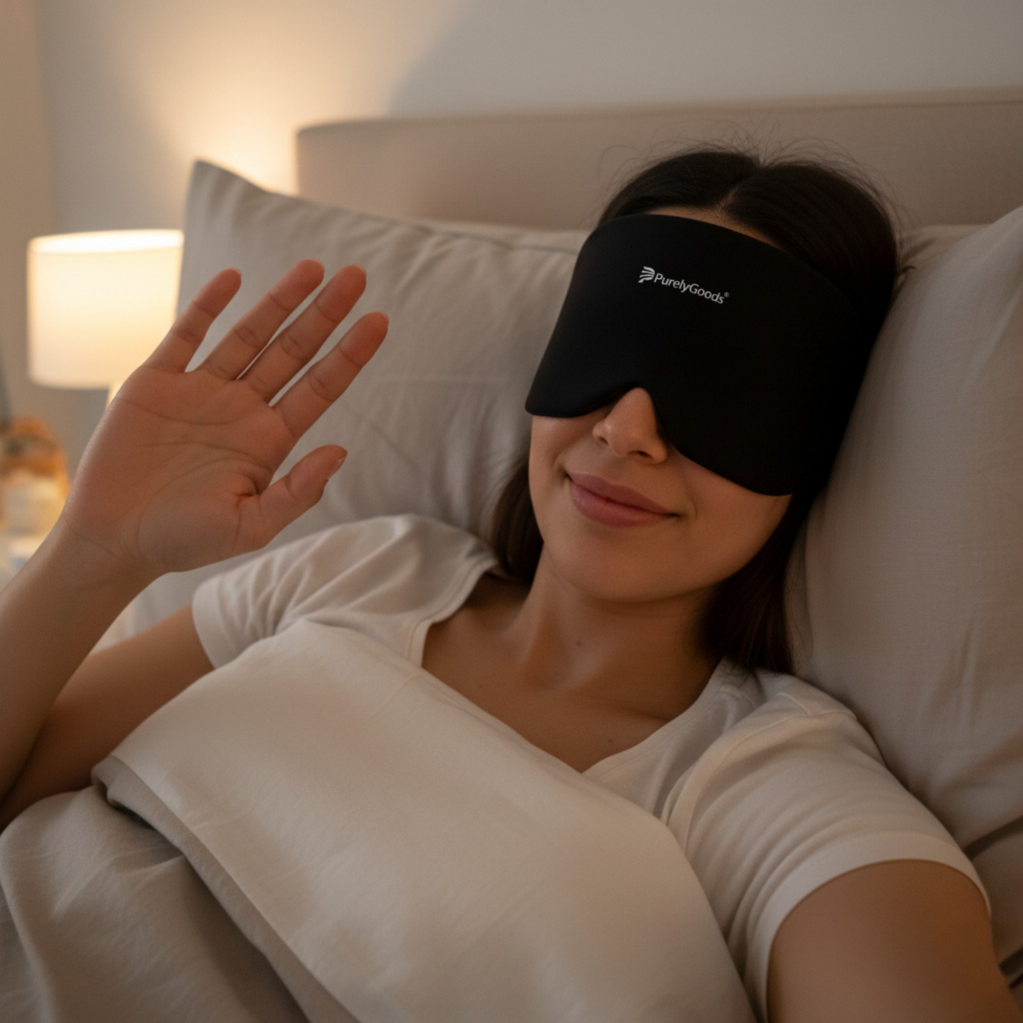 Vrouw ligt ontspannen in bed met een zwart PurelyGoods migraine masker en zwaait rustig naar de camera.