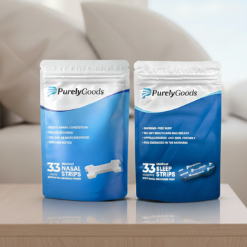 Twee witte en blauwe zakjes met het PurelyGoods-logo staan op een houten oppervlak. Het linkerzakje is voor "Nasal Strips" en toont drie witte neusstrips. Het rechterzakje is voor "Sleep Strips" en toont een blauwe mondtape. Op de achtergrond is een onscherpe witte bank te zien.